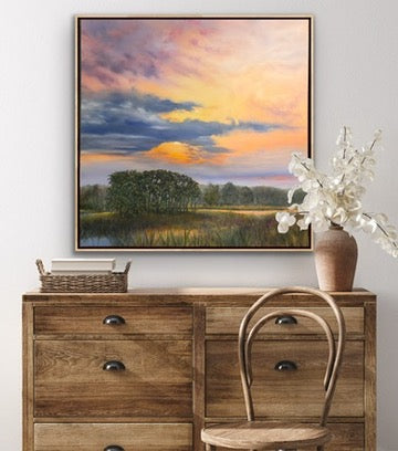 A framed landscape painting of a sunset over a marshland with trees and clouds, displayed on a light gray wall above a rustic wooden dresser with a chair and a vase of white flowers.