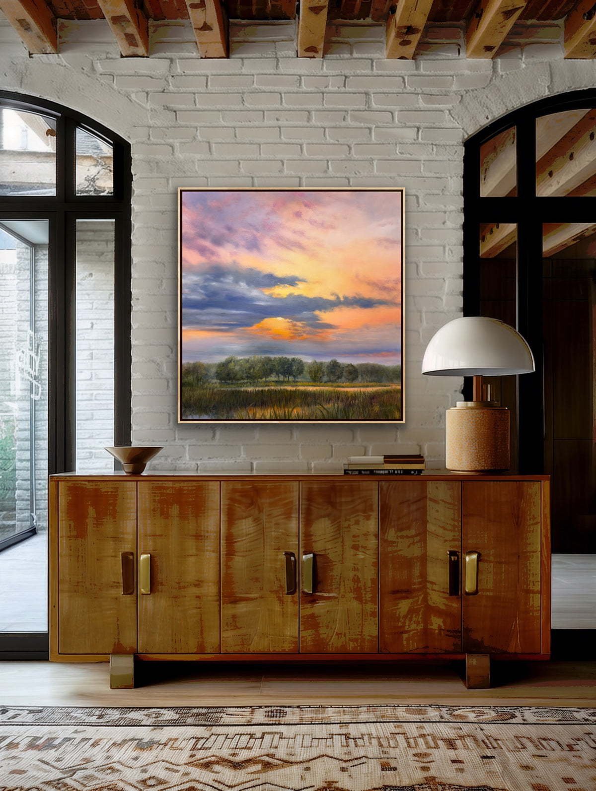 Orange colored sky filled with clouds with the sun peeking through shining on the wetland and trees below. framed hanging on a brick wall above a wooden buffet.